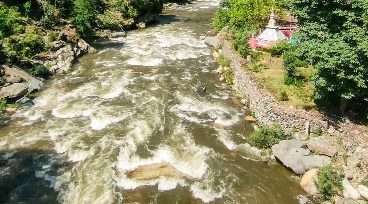 The Tirthan river rushing over boulders beside a stone embankment and a small white temple, just before Banjar