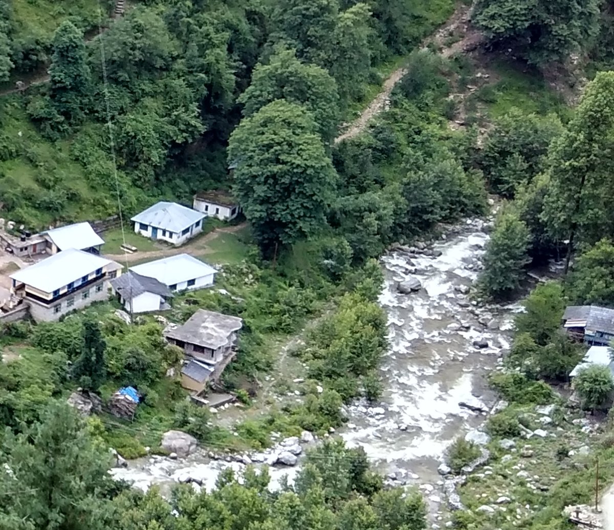 A fast, rocky mountain stream running past village houses and green slopes at Bathad in the Tirthan valley