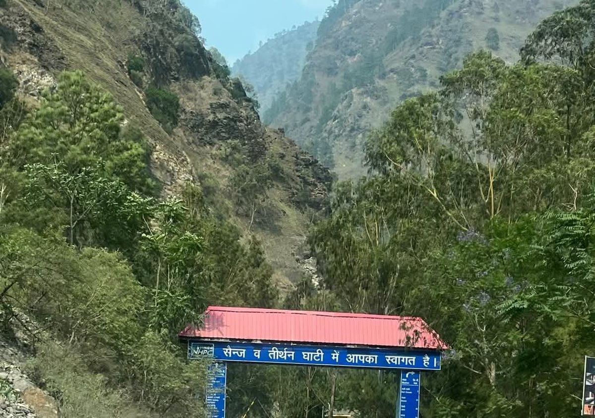 A blue roadside welcome arch at Larji reading, in Hindi, welcome to the Sainj and Tirthan valleys, with forested gorge slopes behind