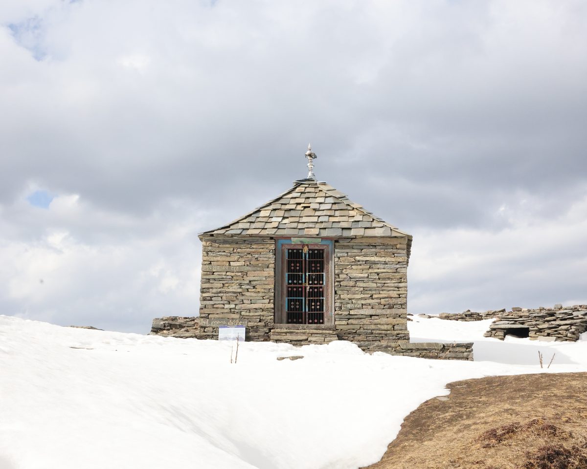 A small stone shrine with a slate roof standing in deep snow among low ruined walls at Raghupur Fort