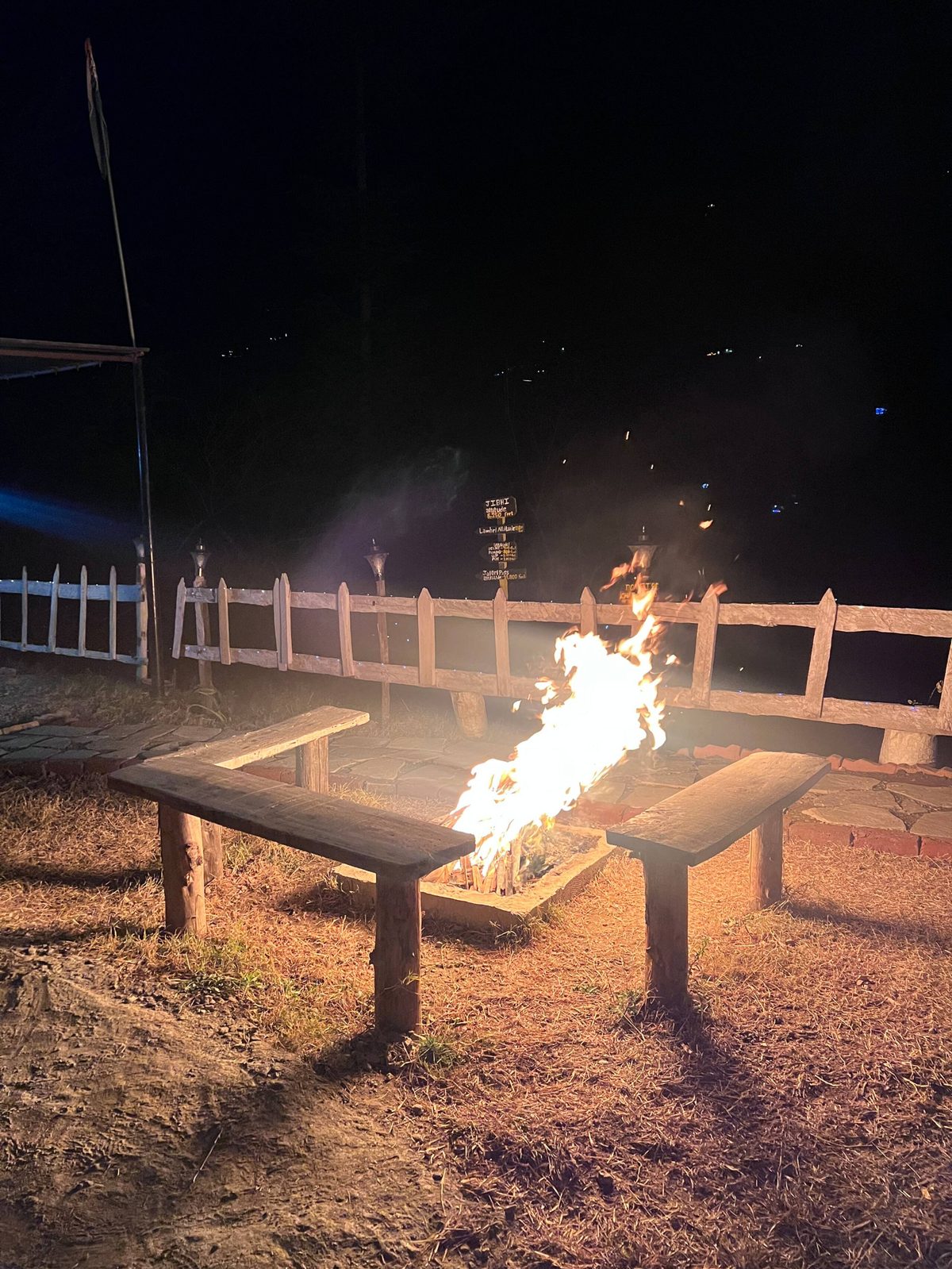 A bonfire burning in a stone fire pit between two wooden benches on the lawn at night at Peaks N Valley Stays in Chhet, near Jibhi