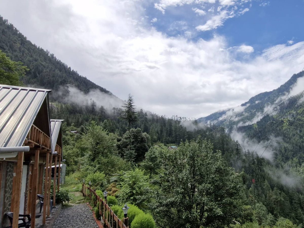 Mist rising over a green forested valley beside the Mudhouse cottages and a gravel path at Peaks N Valley Stays in Chhet, near Jibhi