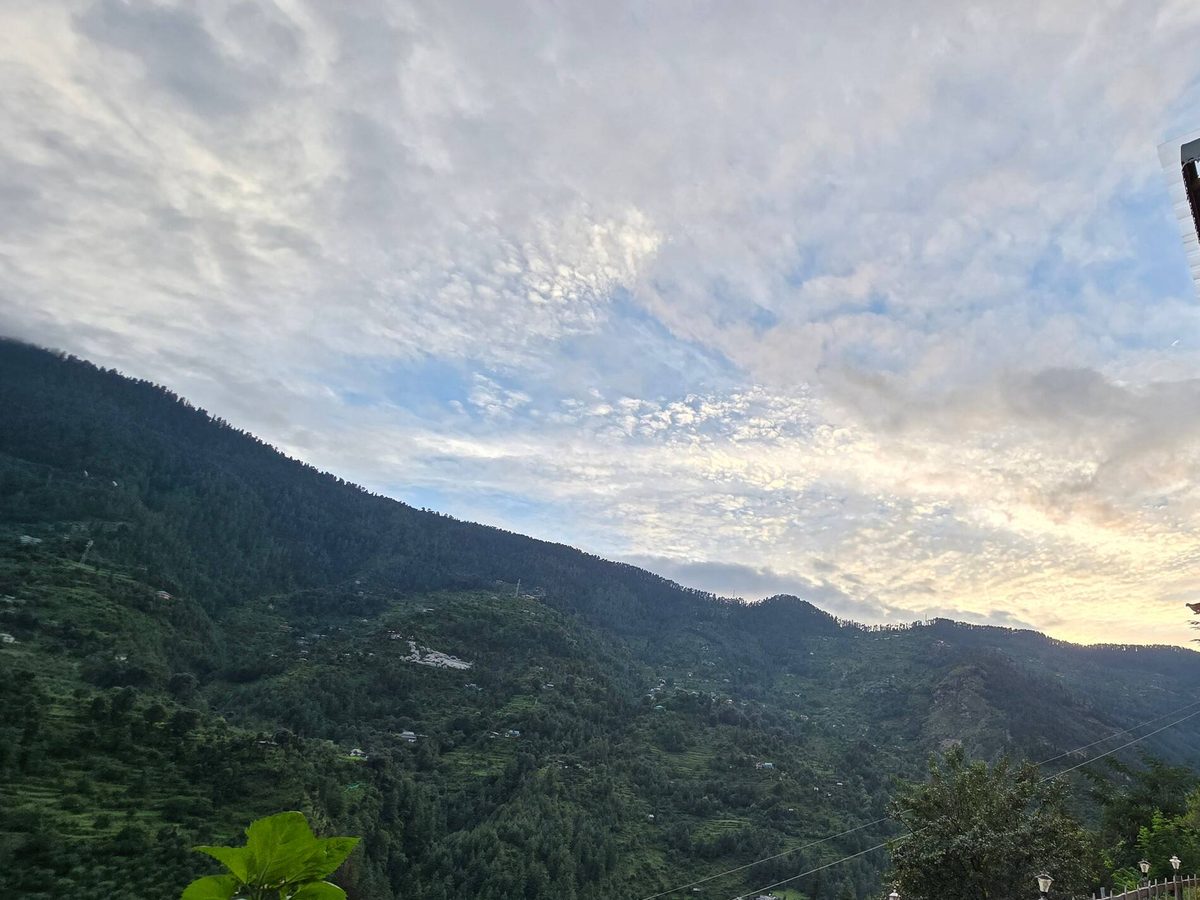 Green forested valley under a cloudy sky beside the Glass House, with a stone path and fence at Peaks N Valley Stays near Jibhi